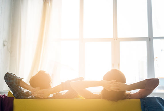 două persoane care se bucură de beneficile masajului stând relaxați pe o canapea cu fata catre soarele care intra pe fereastra